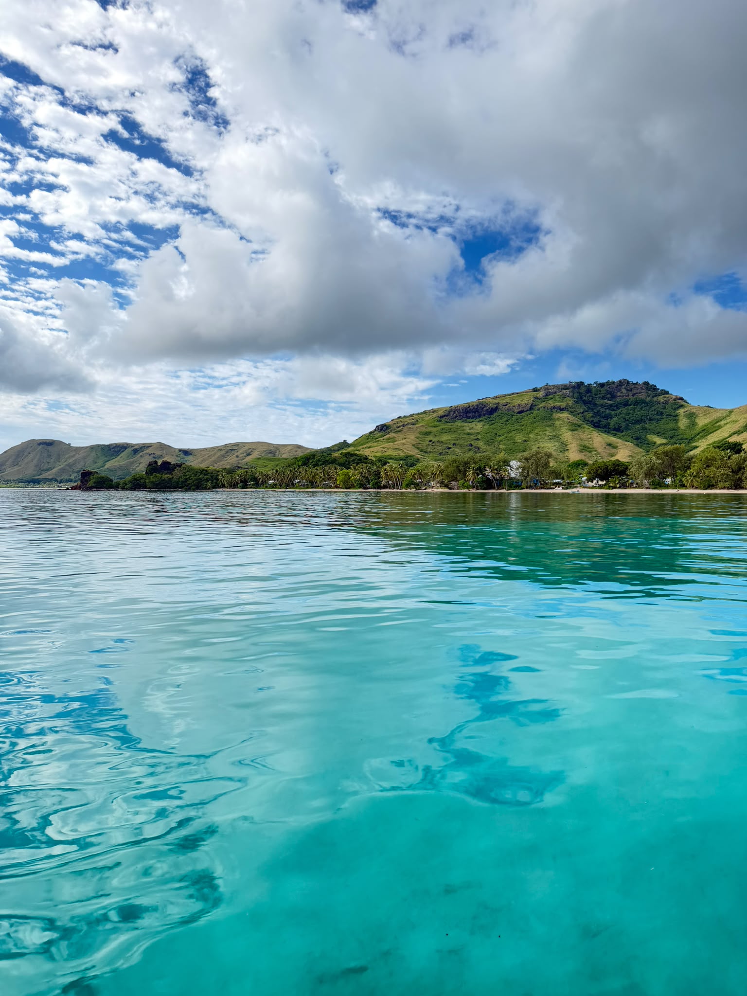 A clear turquoise lagoon meeting green hills beneath a wide, cloud-filled sky in Fiji